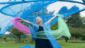 A woman in a blue cardigan dances with pink and green chiffon ribbons outdoors